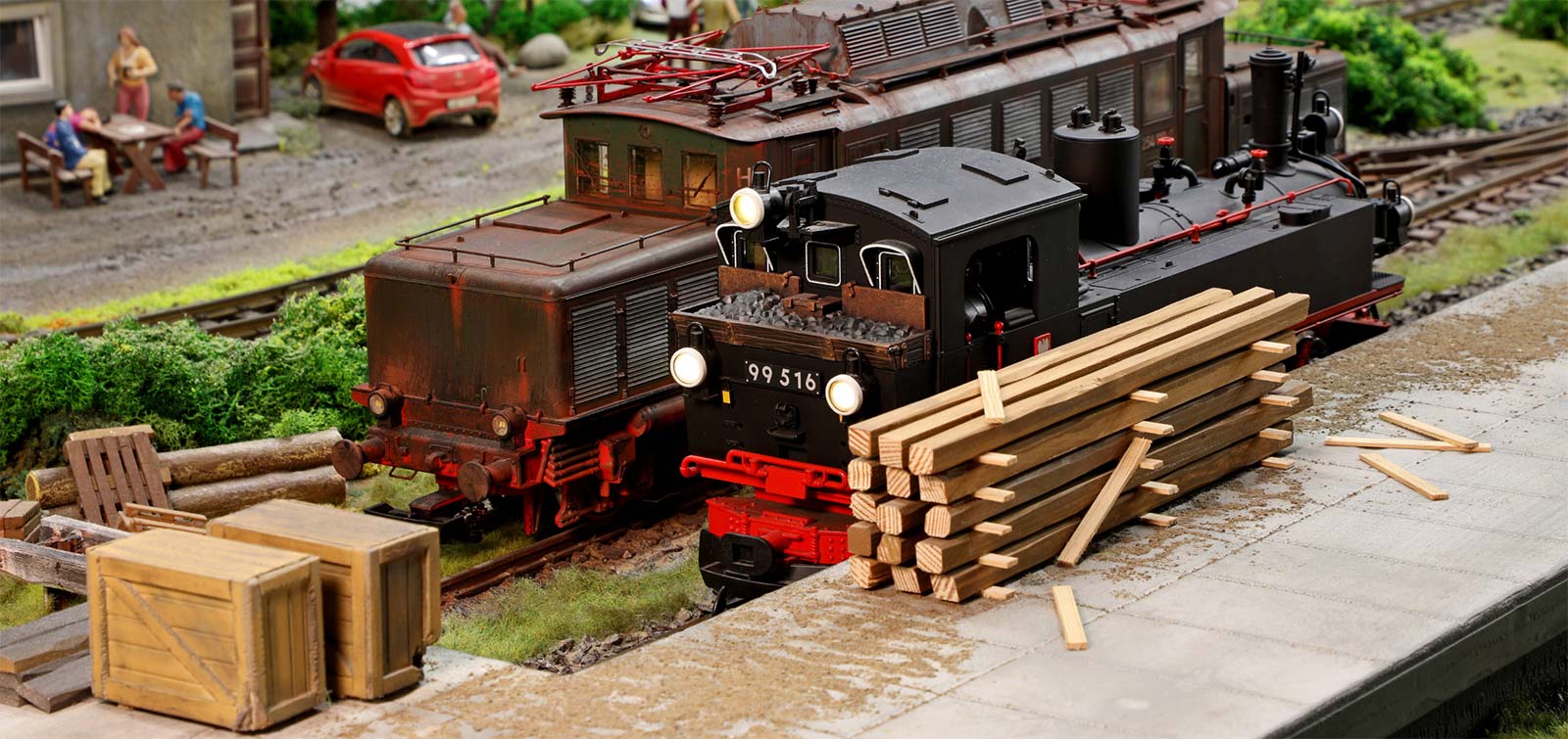 Zdjęcie przedstawia miniaturowy parowóz i elektrowóz oraz peron z rozładowanym drewnem na wystawie Kolejkowo Wrocław